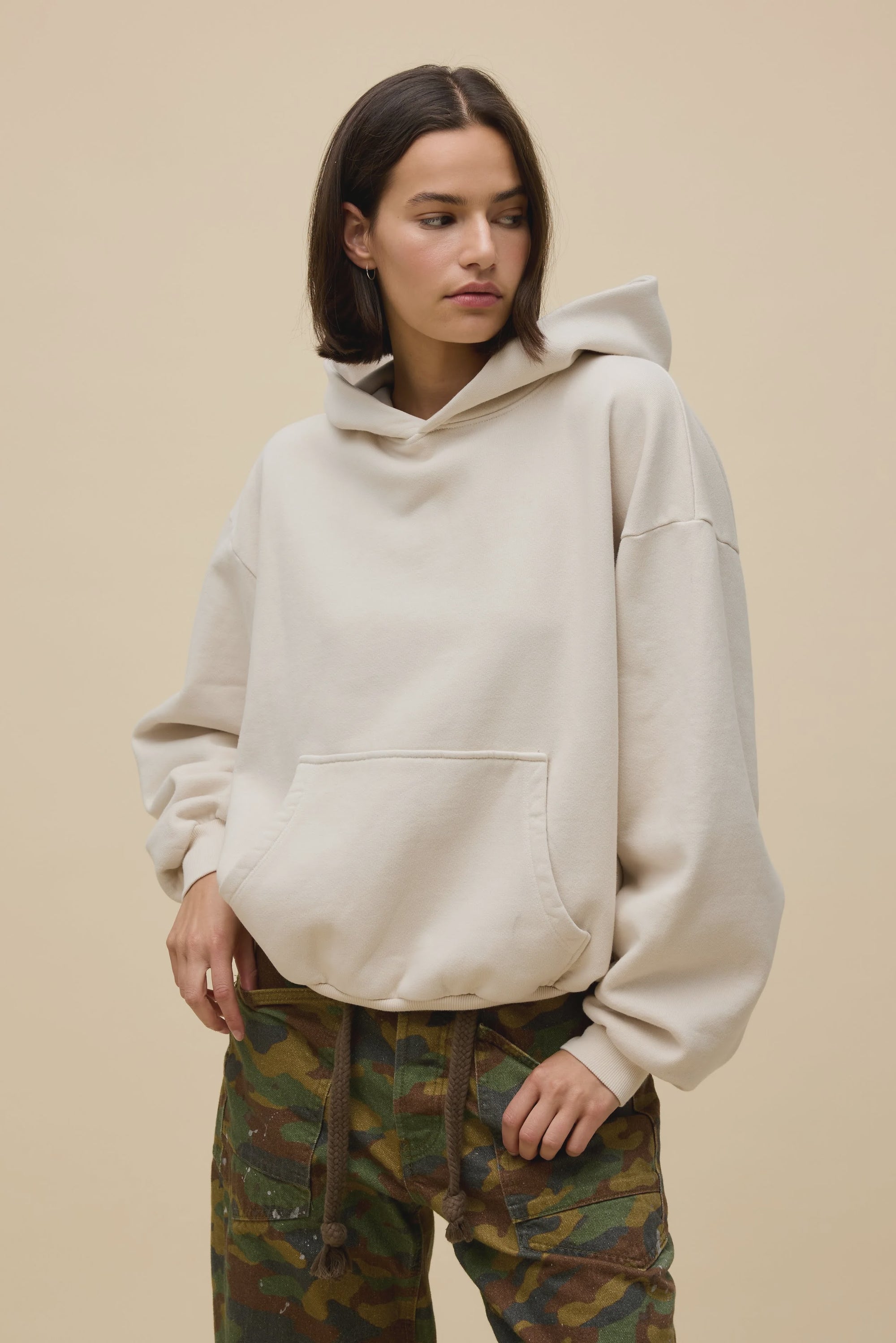A woman models the DayDreamer OS Hoodie, an oversized beige style, paired with camouflage pants. She stands against a plain beige background, gazing slightly left with one hand in her pocket.