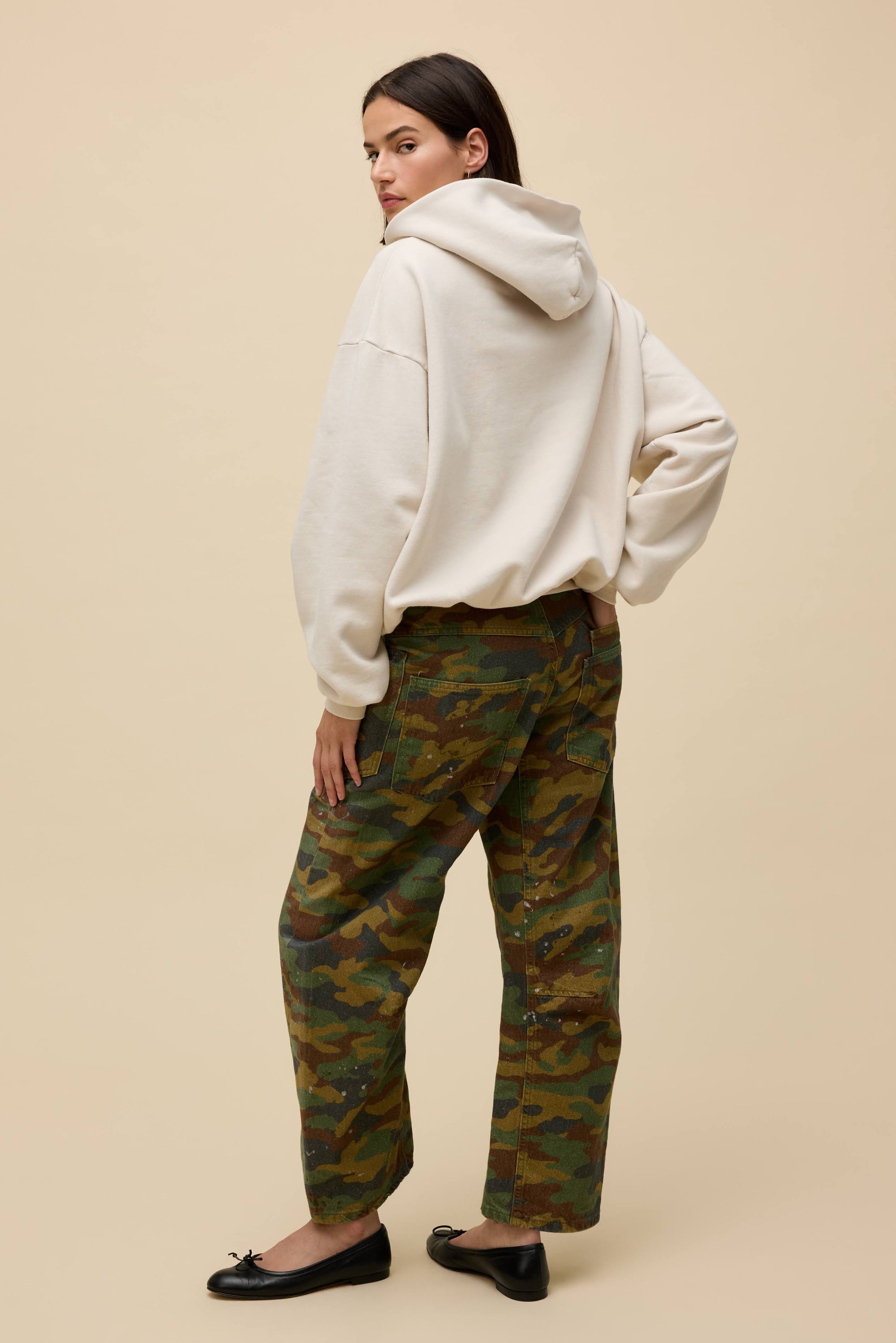 A person stands facing away against a beige background, wearing the DayDreamer OS Hoodie in cream, paired with green camouflage pants and black flats. Their head is turned slightly to the side.