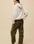 A person stands facing away against a beige background, wearing the DayDreamer OS Hoodie in cream, paired with green camouflage pants and black flats. Their head is turned slightly to the side.