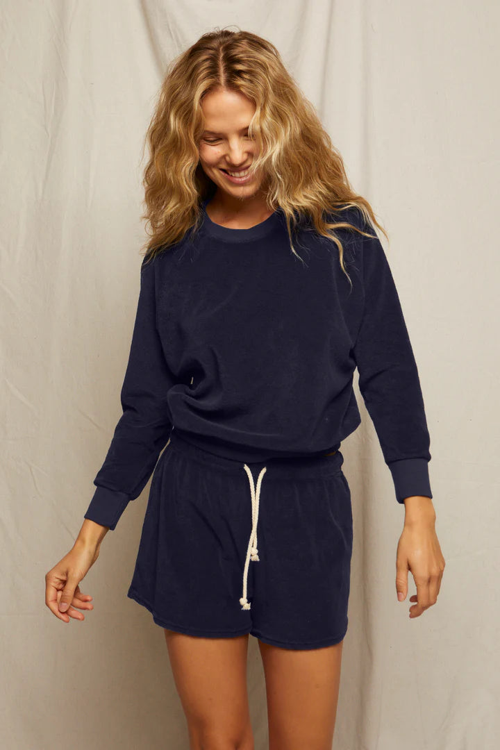 A woman with wavy blonde hair smiles while wearing the Perfectwhitetee Saylor Loop Terry Sweatshirt in navy blue and matching shorts with a white drawstring, standing before a light background.