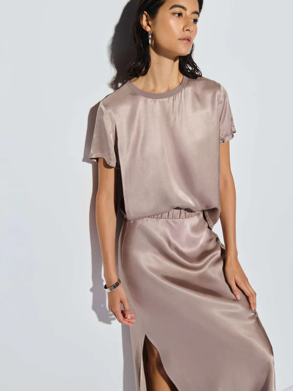 A woman wearing the elegant Marie Boxy Crop by Nation LTD stands against a neutral background. The silky, light beige top pairs perfectly with her lightweight skirt, which features a chic slit. With hoop earrings completing her look, she gazes to her left while resting her right hand on her hip.