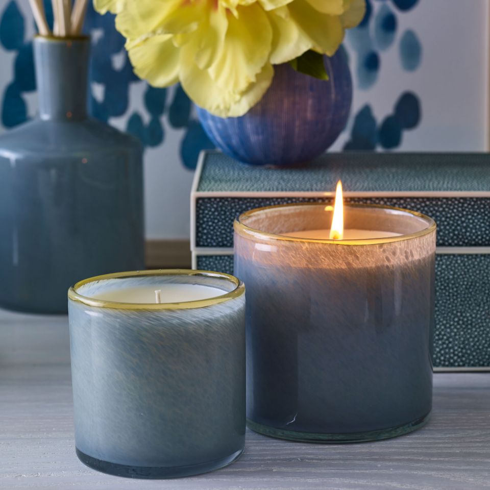 Two blue Beach House candles by Lafco NY in hand-blown glass sit side by side on a table; one glows while the other is unlit. A yellow flower in a vase and a decorative book or box evoke the Sea & Dune-inspired ambiance.