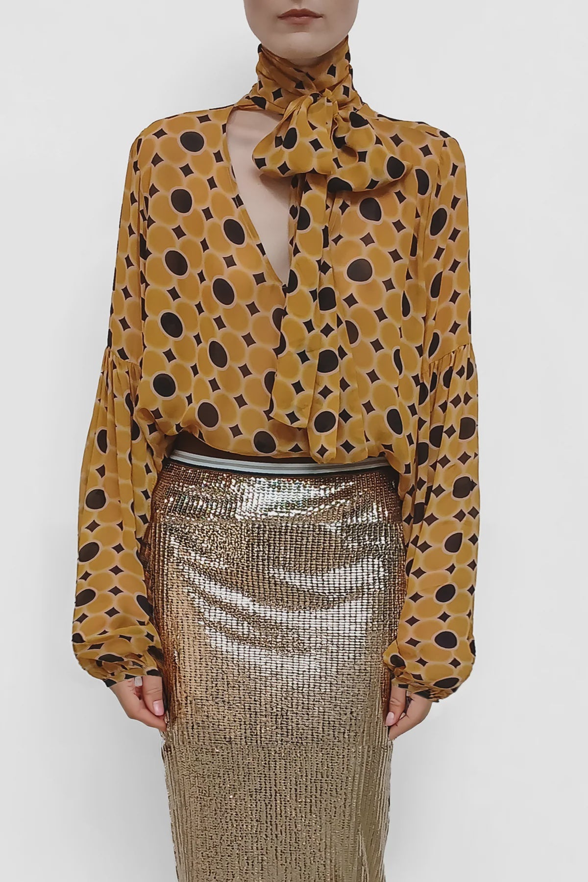 A person wears the Serotonin Blouse by Le Superbe—a yellow polka dot top with dramatic sleeves and a high neck bow—paired with a metallic gold skirt. The cropped photo shows only their upper body and lower face.