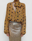 A person wears the Serotonin Blouse by Le Superbe—a yellow polka dot top with dramatic sleeves and a high neck bow—paired with a metallic gold skirt. The cropped photo shows only their upper body and lower face.