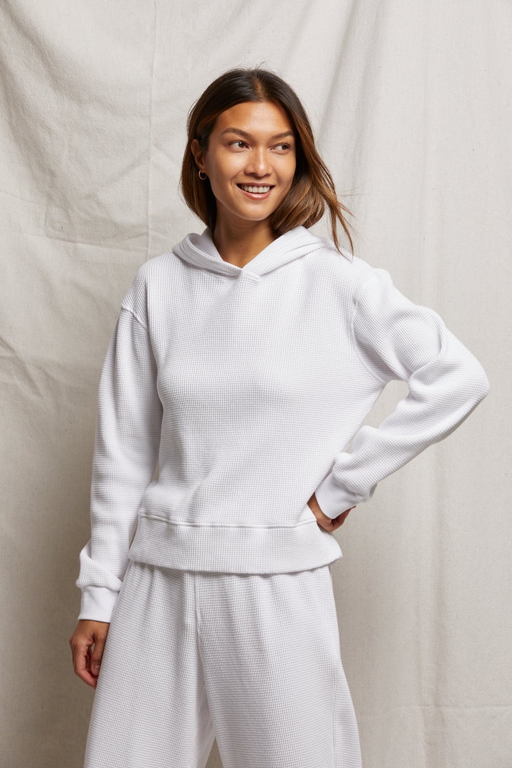 A person dresses in a Perfectwhitetee azalia waffle hoodie and matching pants, posing stylishly against a neutral fabric backdrop with a smile and one hand on their hip, embodying effortless travel lounge fashion.