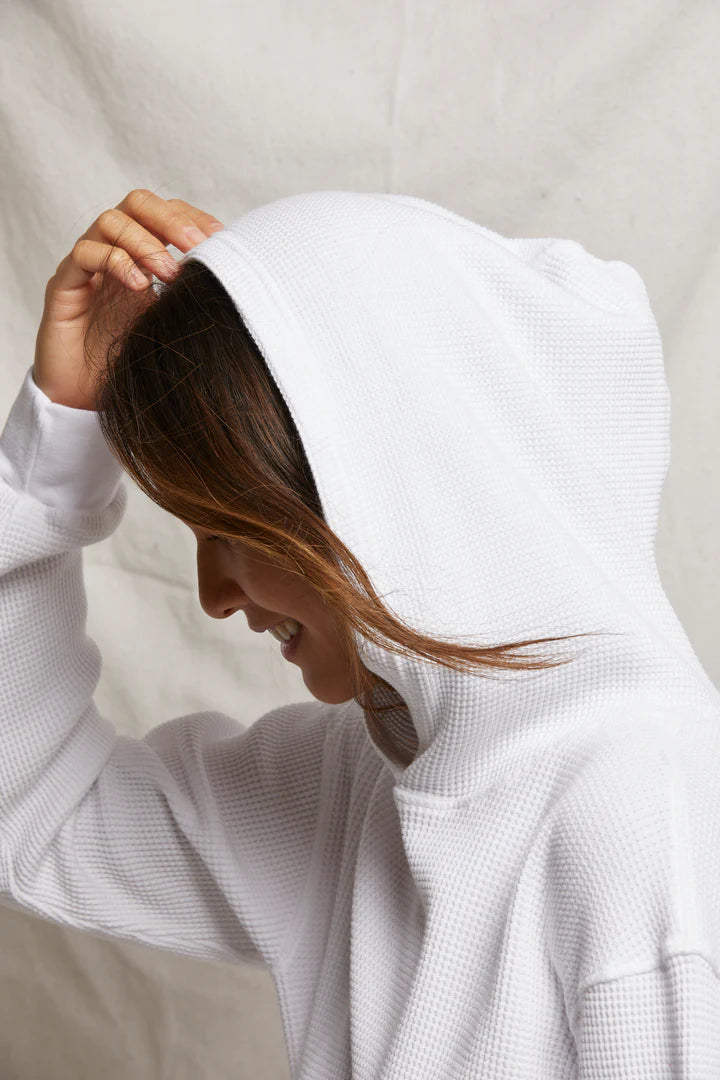 A person smiles while touching the hood of a "Perfectwhitetee" azalia waffle hoodie. The neutral, light background highlights an effortless style, ideal for travel lounging.