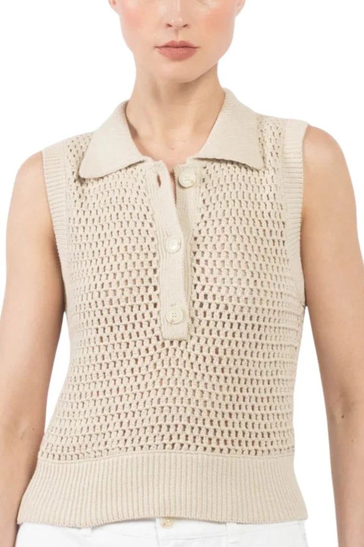 A woman wears FRAME’s The Polo Vest—a sleeveless, beige, crochet-style knit wool top with a collar and three front buttons—paired with white pants for a chic monochrome look.