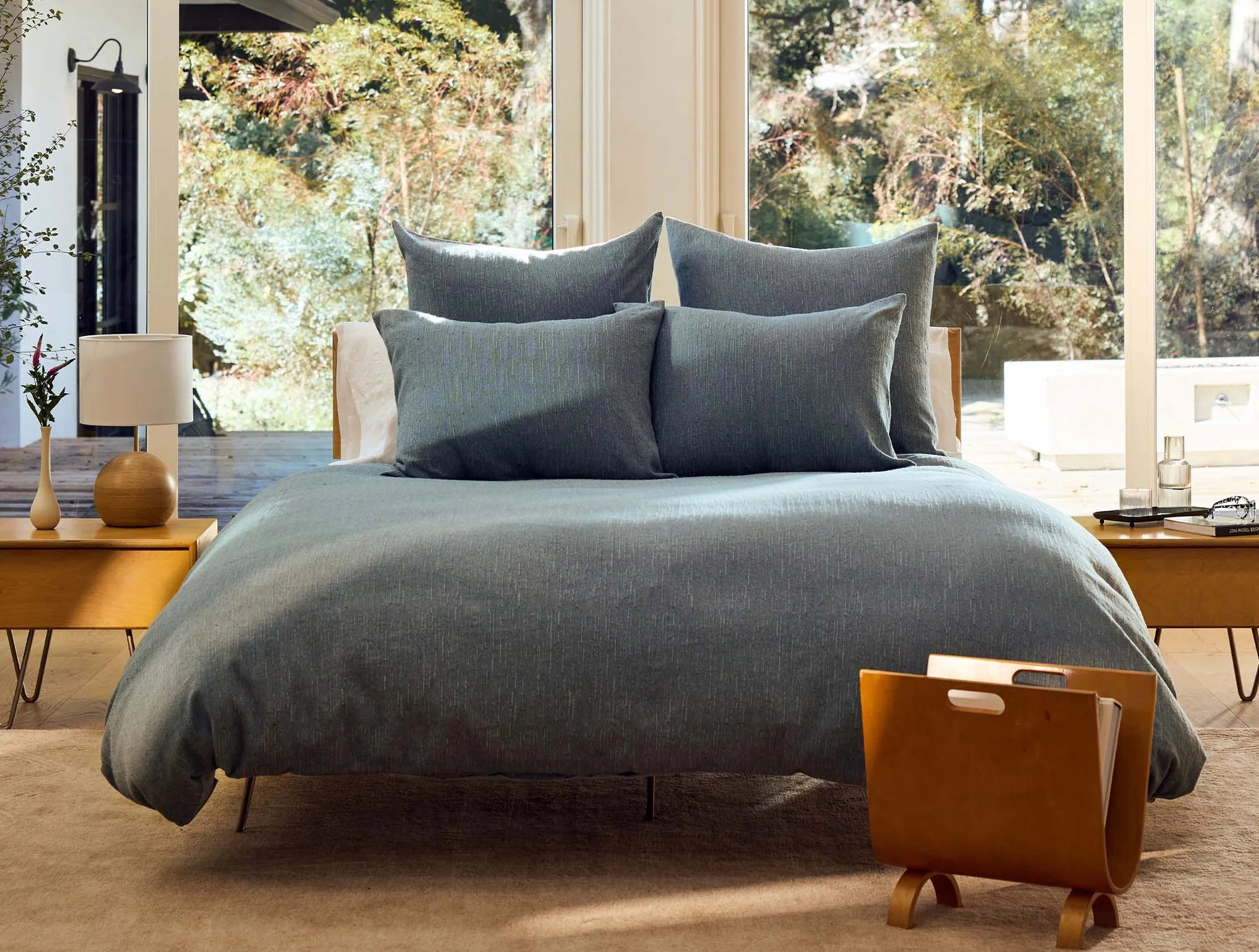 A sunlit bedroom features a neatly made bed with the Coyuchi Inc Toro Canyon Org. Duvet Cover in Marine King, four pillows, wooden side tables, a lamp, and a magazine holder by large windows overlooking trees.
