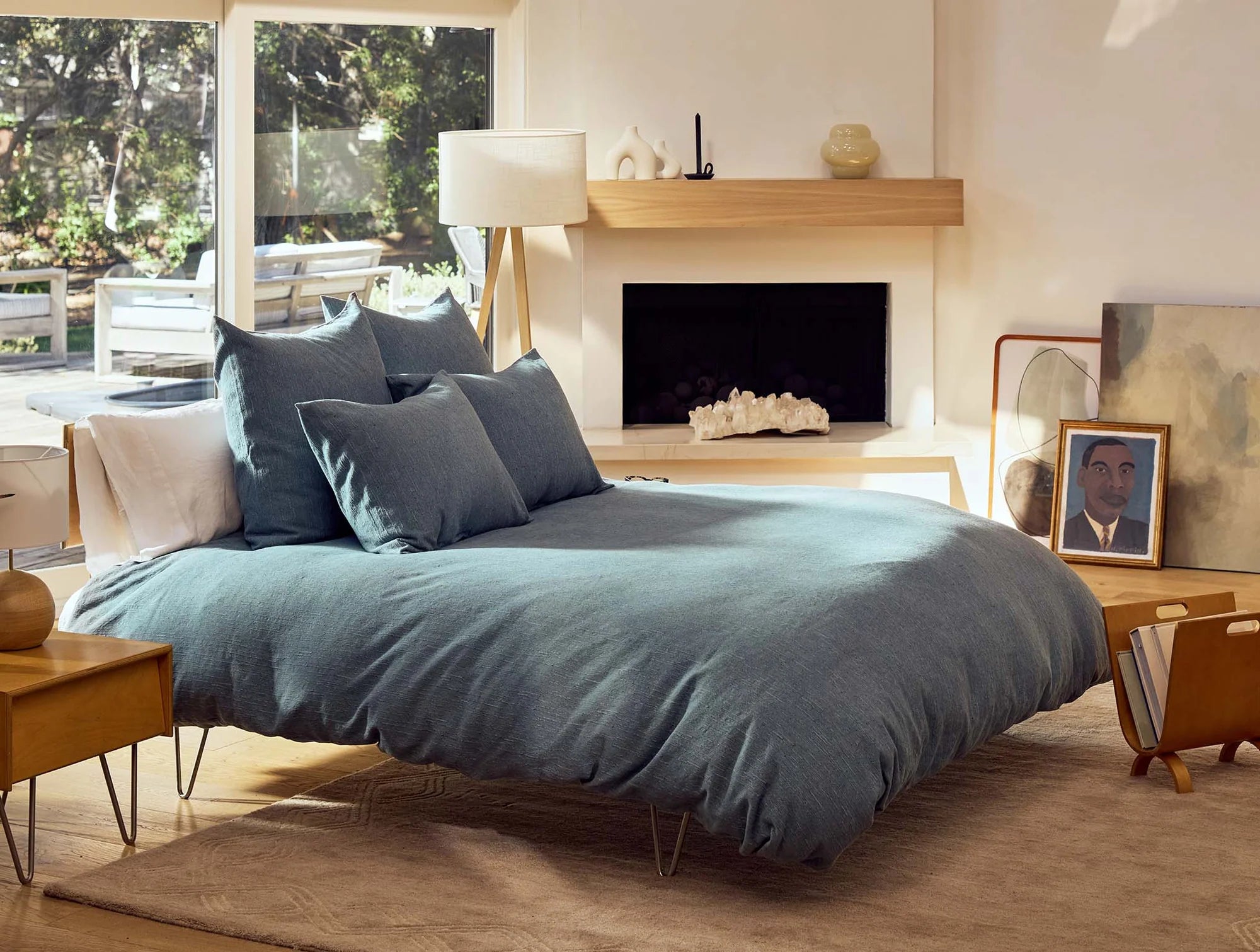 A cozy bedroom with a large bed dressed in blue bedding and the Toro Canyon Org, Euro Sham in Marine by Coyuchi Inc, surrounded by mid-century modern furniture, a lamp, textile-inspired wall art, and large windows filled with natural light.