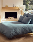 A sunlit bedroom with a modern blue bed, large pillows, wooden furniture, a fireplace, a portrait on the floor, and big windows overlooking greenery features the Coyuchi Toro Canyon Org. Duvet Cover Marine Queen.
