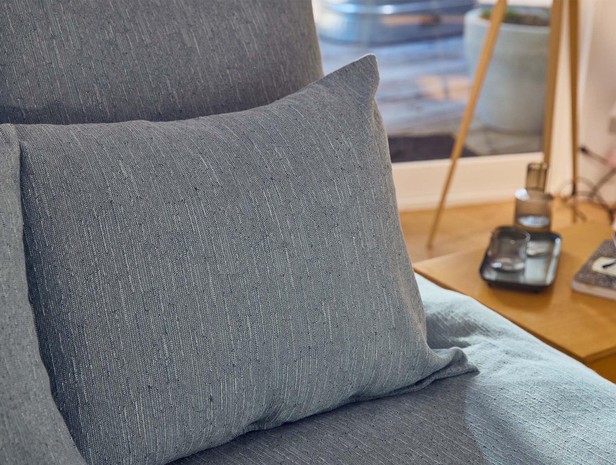 A close-up of the Toro Canyon Org, Euro Sham Marine by Coyuchi Inc on a bed with matching bedding, beside a wooden nightstand holding a tray and bottle. Sunlight highlights its mid-century textile artist-inspired weave.