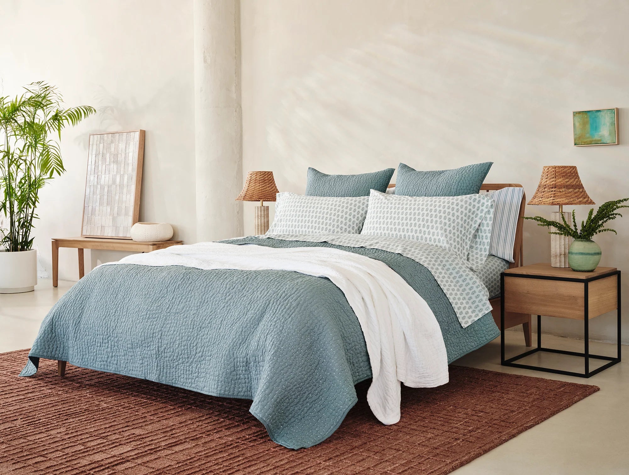 A modern bedroom with a bed covered in blue and white 100% organic cotton bedding, Coyuchi Org Percale Pillowcase Dew Chrysanthemum S/2 Queen, two wooden nightstands with woven lamps, brown rug, potted plant, and neutral wall decor in a bright space.