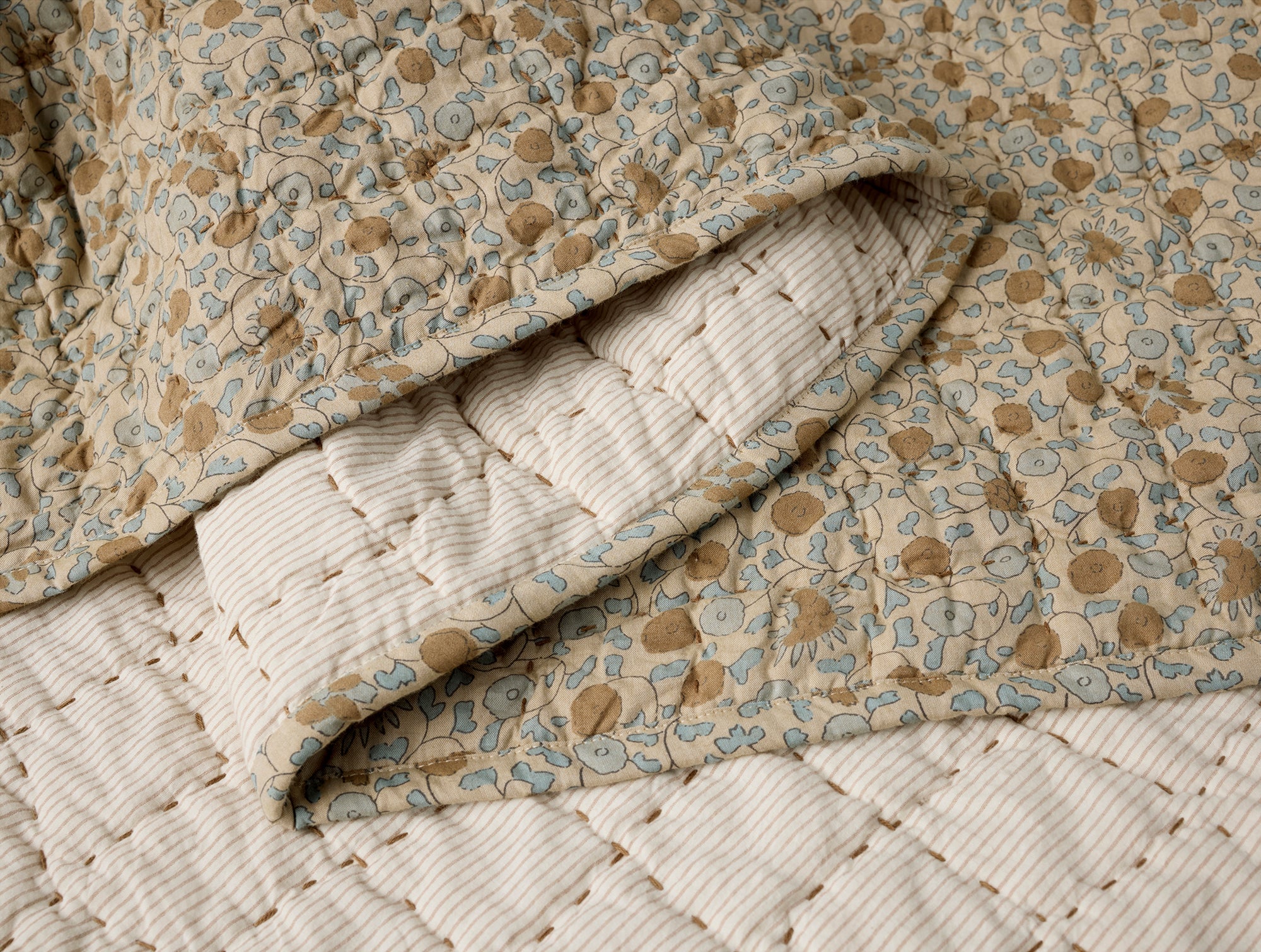 A close-up of the Blossom Hand Stitched Quilt by Coyuchi Inc shows its beige, blue, and brown floral design, with a cream diamond-stitched underside—crafted from organic cotton for comfort and style.