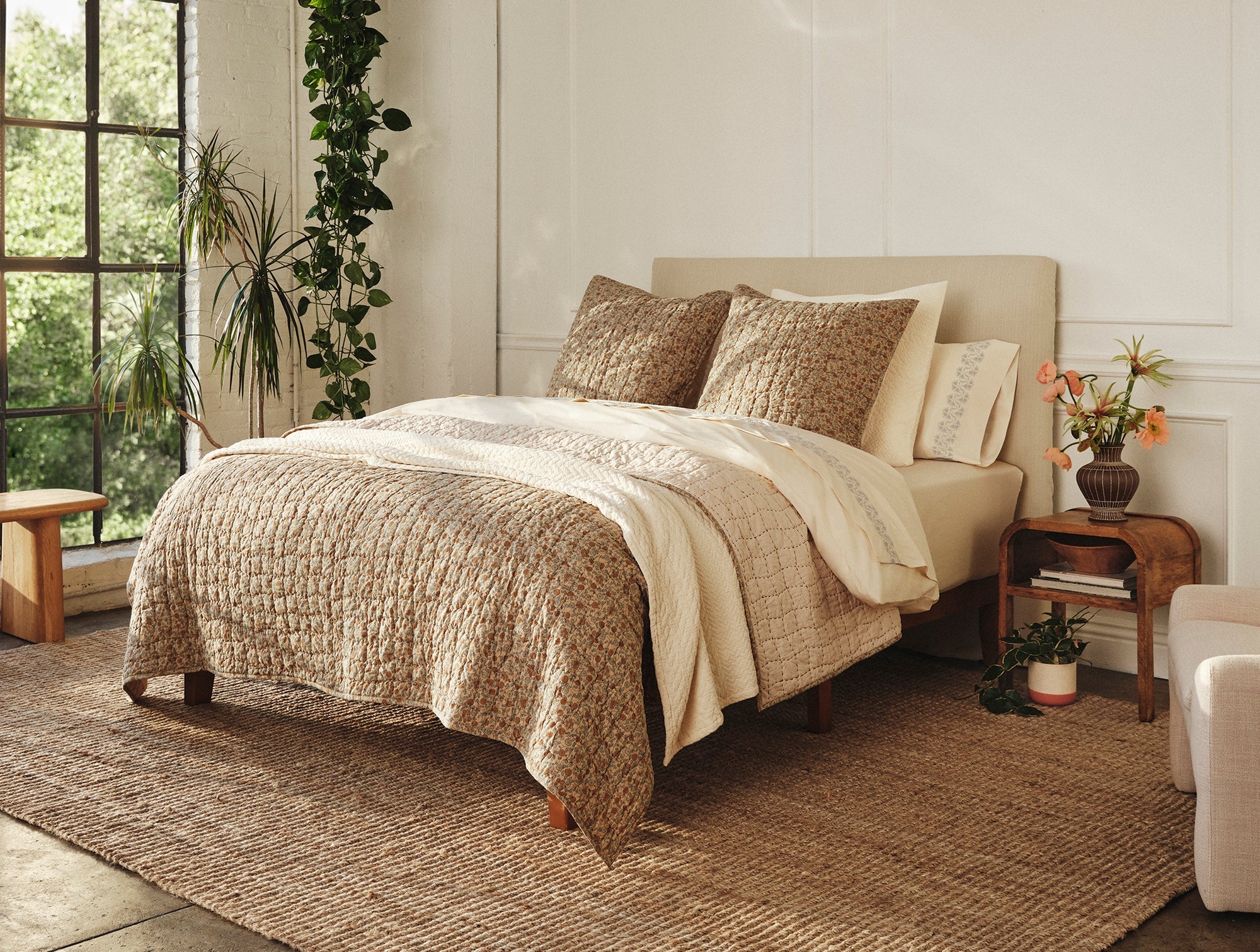 A cozy bedroom with a neatly made bed featuring the Blossom Hand Stitched Quilt by Coyuchi Inc, pillows, a cream headboard, plants, wooden furniture, a large window with greenery outside, and a woven rug on the floor.