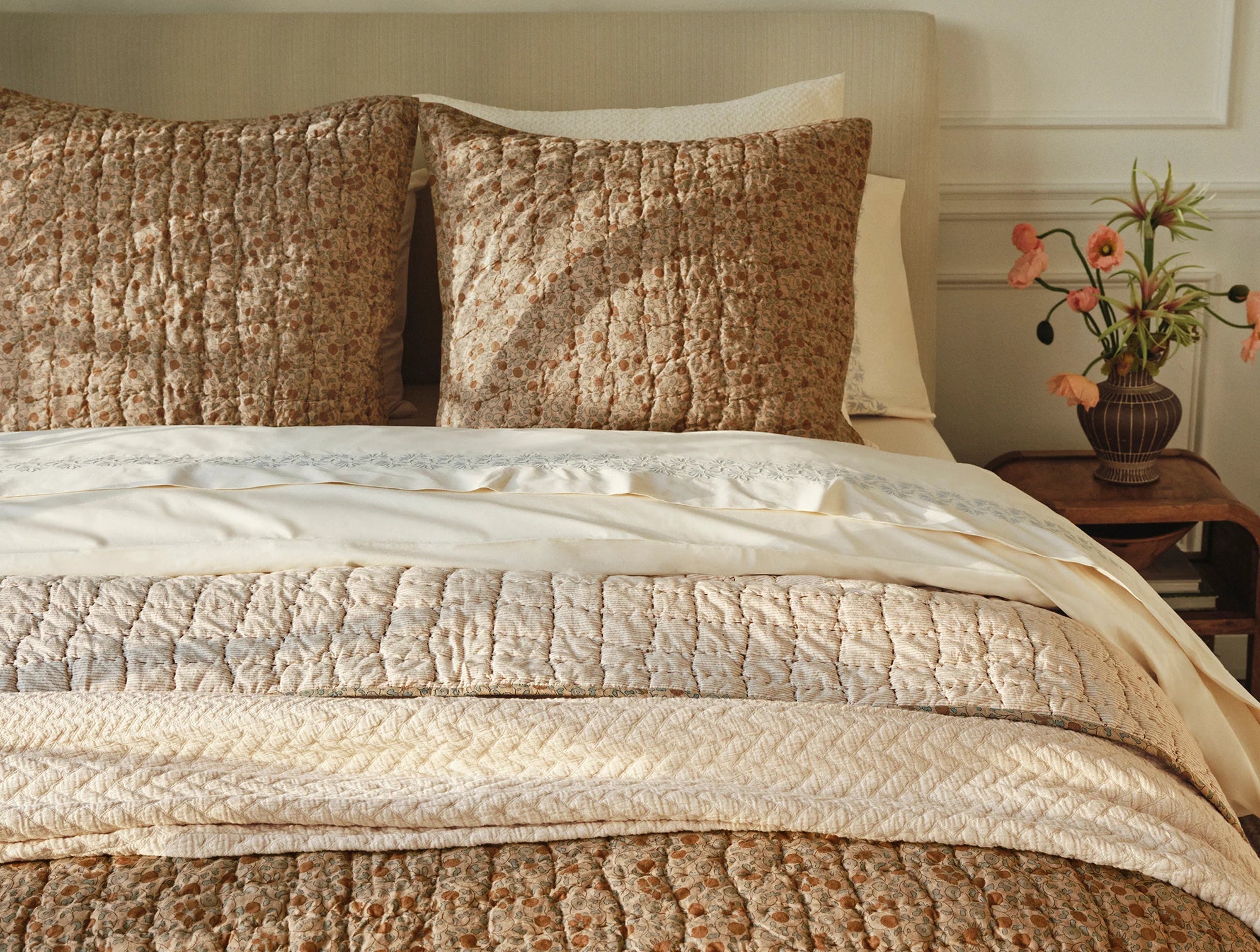 A softly lit, elegant bedroom features a neatly made bed with Coyuchi Inc’s Blossom Hand Stitched Euro Sham and textured brown and beige pillows, beside a wooden nightstand topped with a dark vase of orange and pink flowers.
