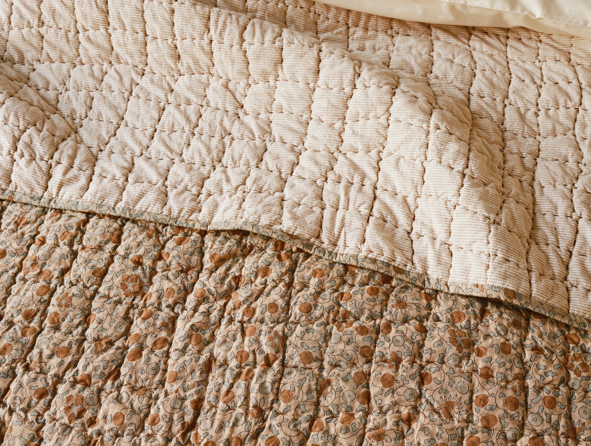 A close-up of a bed with two quilts: on top is Coyuchi Inc’s Blossom Hand Stitched Quilt in light beige with a grid pattern, while the bottom quilt shows a floral design with orange and green accents.