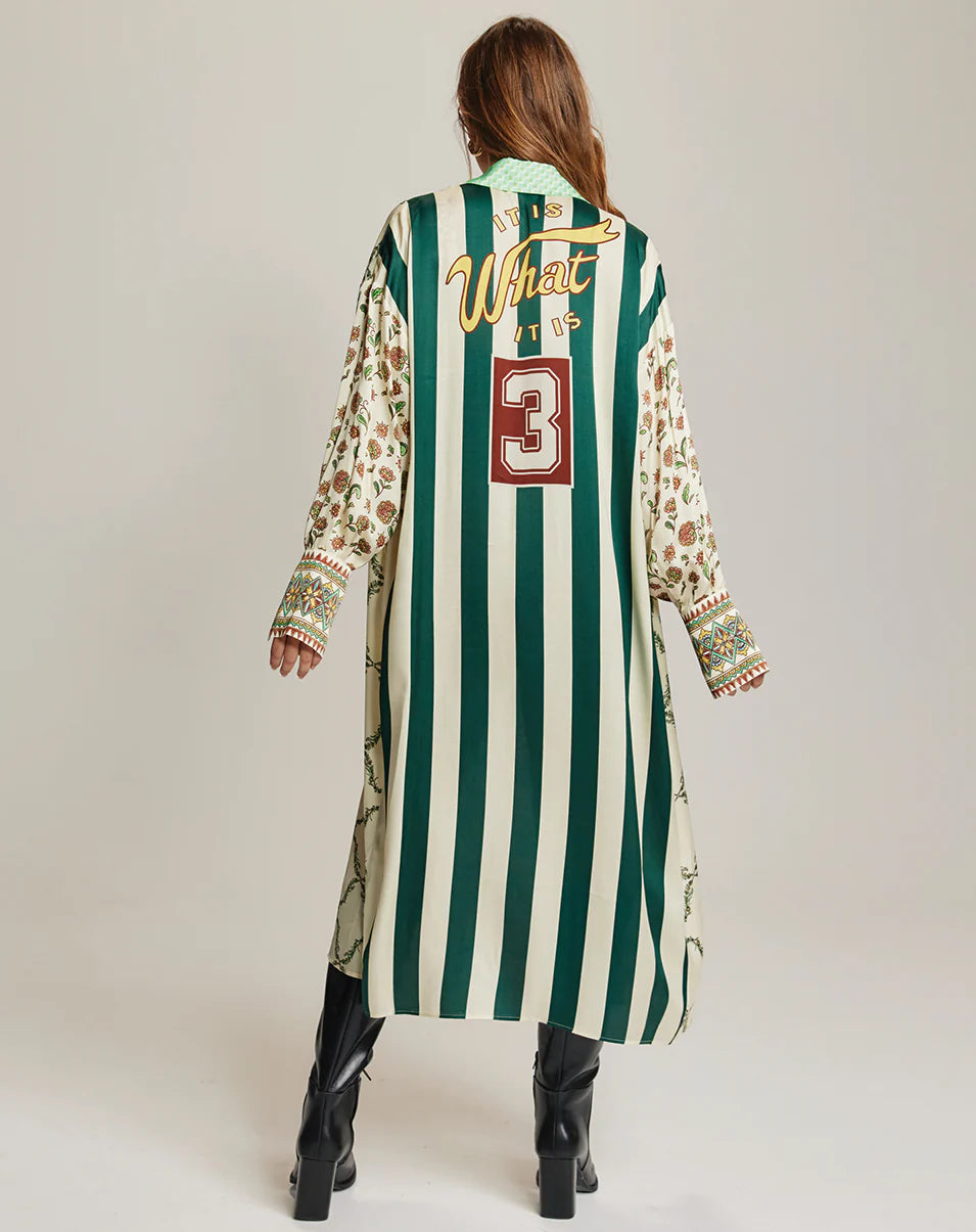 A woman faces away, wearing the Me369 Athena Collar Button Up dress with green and white stripes, floral sleeves, embroidery, and a button-up collar. "What it is" and a large red 3 are on the back. She pairs it with black high-heeled boots.