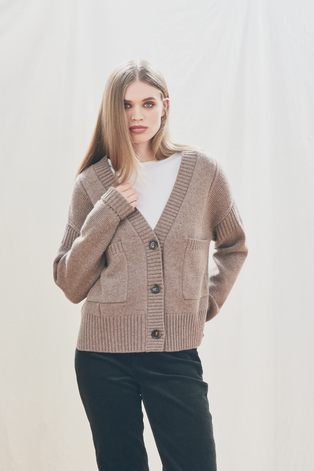 A woman with straight, light brown hair wears the Jumper 1 2 3 4/CR2 Guernsey Cardigan in fawn grey over a white shirt and dark pants, standing against a light, neutral background.
