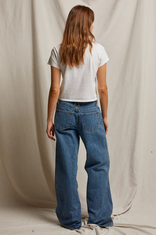 A woman with long brown hair faces away, wearing the Perfectwhitetee Springsteen Baby Tee in white and loose blue jeans, set against a beige fabric backdrop.