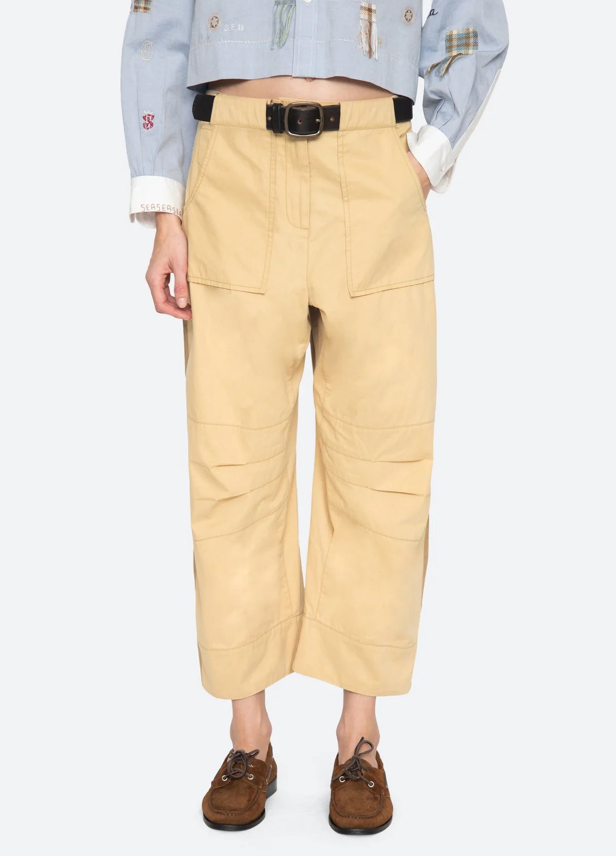 The lower half of a person is shown wearing Sea New York’s Andrea WorkWear Pants—wide-leg, cropped, tan trousers with large front pockets—styled with a black belt, brown shoes, and a light blue shirt with white cuffs.