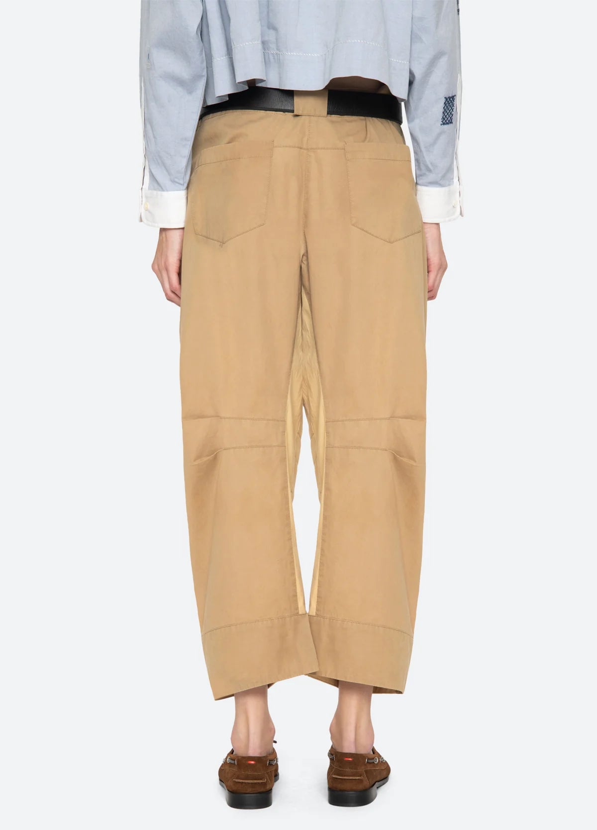 A person stands facing away, wearing Sea New York's Andrea WorkWear Pants in tan with a barrel-leg silhouette and back pockets, paired with brown sandals and a blue long-sleeve shirt for a contemporary statement look.