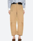A person stands facing away, wearing Sea New York's Andrea WorkWear Pants in tan with a barrel-leg silhouette and back pockets, paired with brown sandals and a blue long-sleeve shirt for a contemporary statement look.