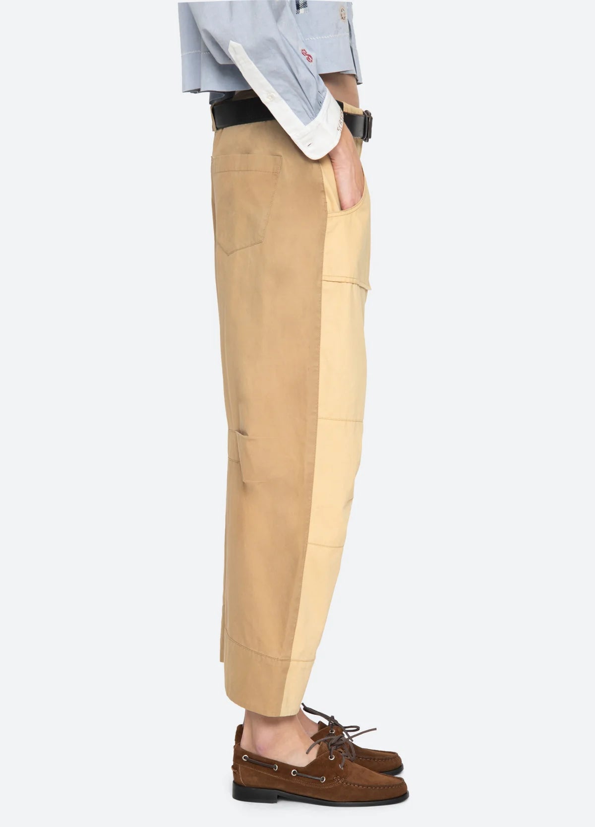 A person stands in profile wearing Sea New York’s Andrea WorkWear Pants—tan patchwork with a barrel-leg silhouette—paired with a light blue button-up and brown boat shoes, left hand in pocket against a plain white background.