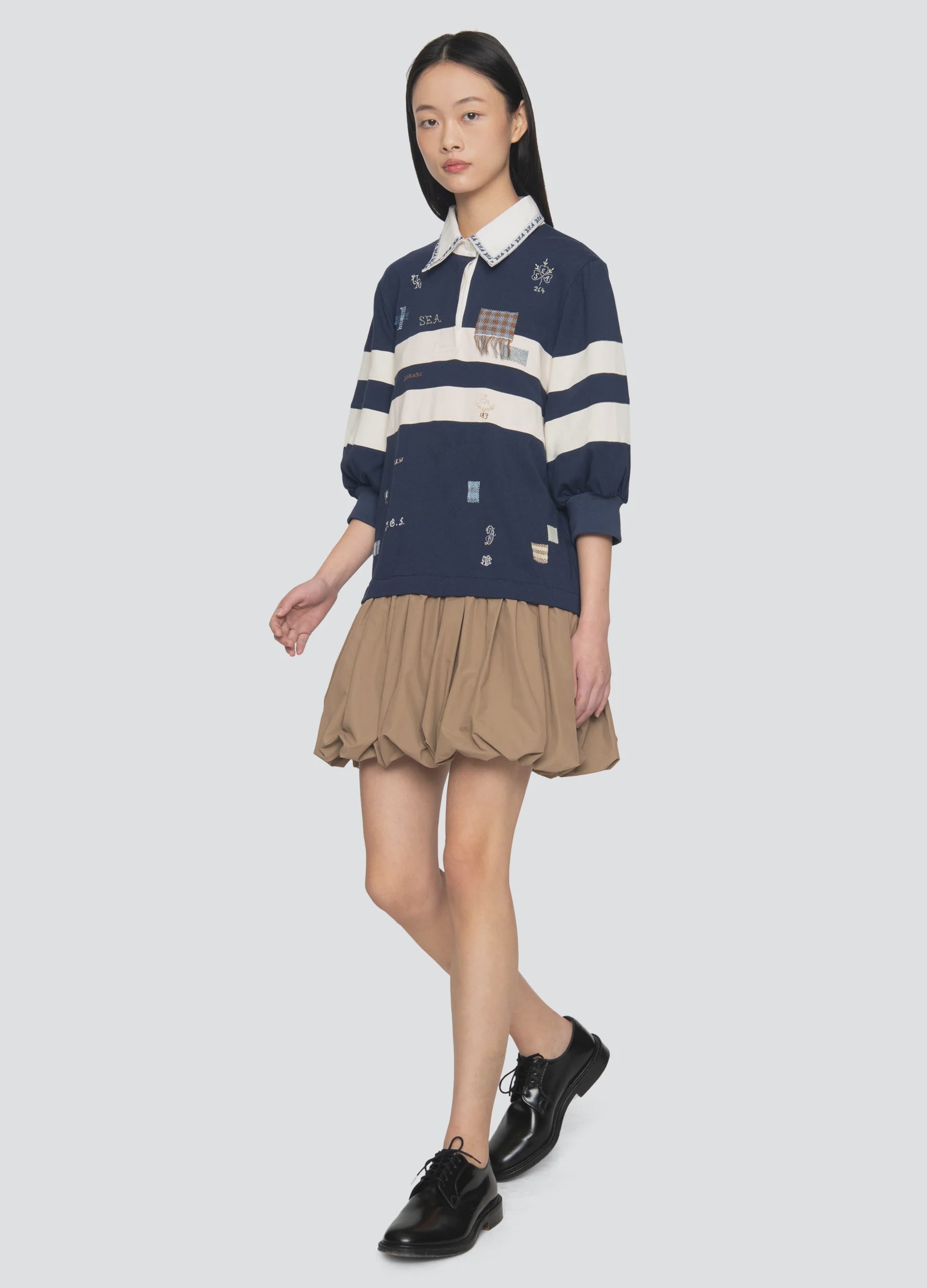 A woman with straight black hair walks against a plain light gray background, wearing the Sea New York Claira Rugby Dress in navy blue with white stripes, paired with black shoes.