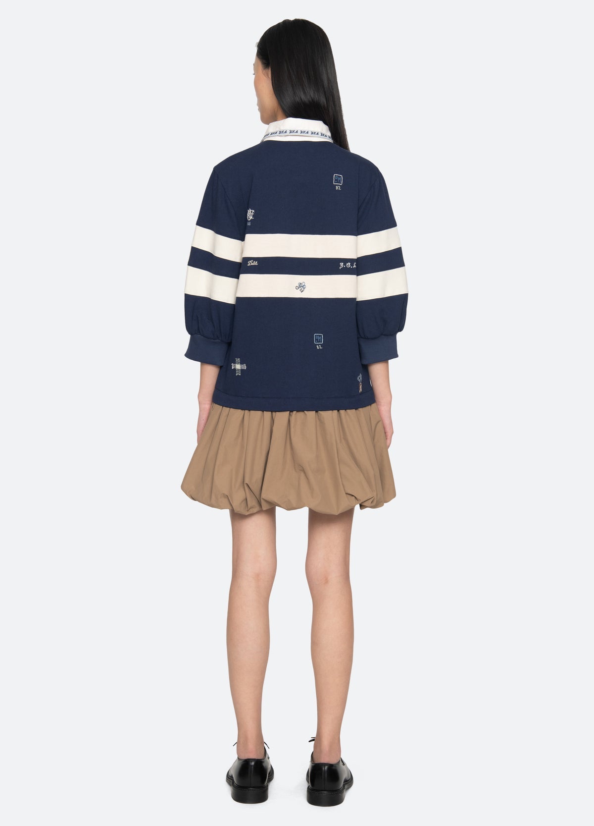 A person with long dark hair is seen from behind, wearing the Sea New York Claira Rugby Dress—a navy blue dress with white horizontal stripes and artisanal graphics—paired with black shoes, standing against a plain white background.