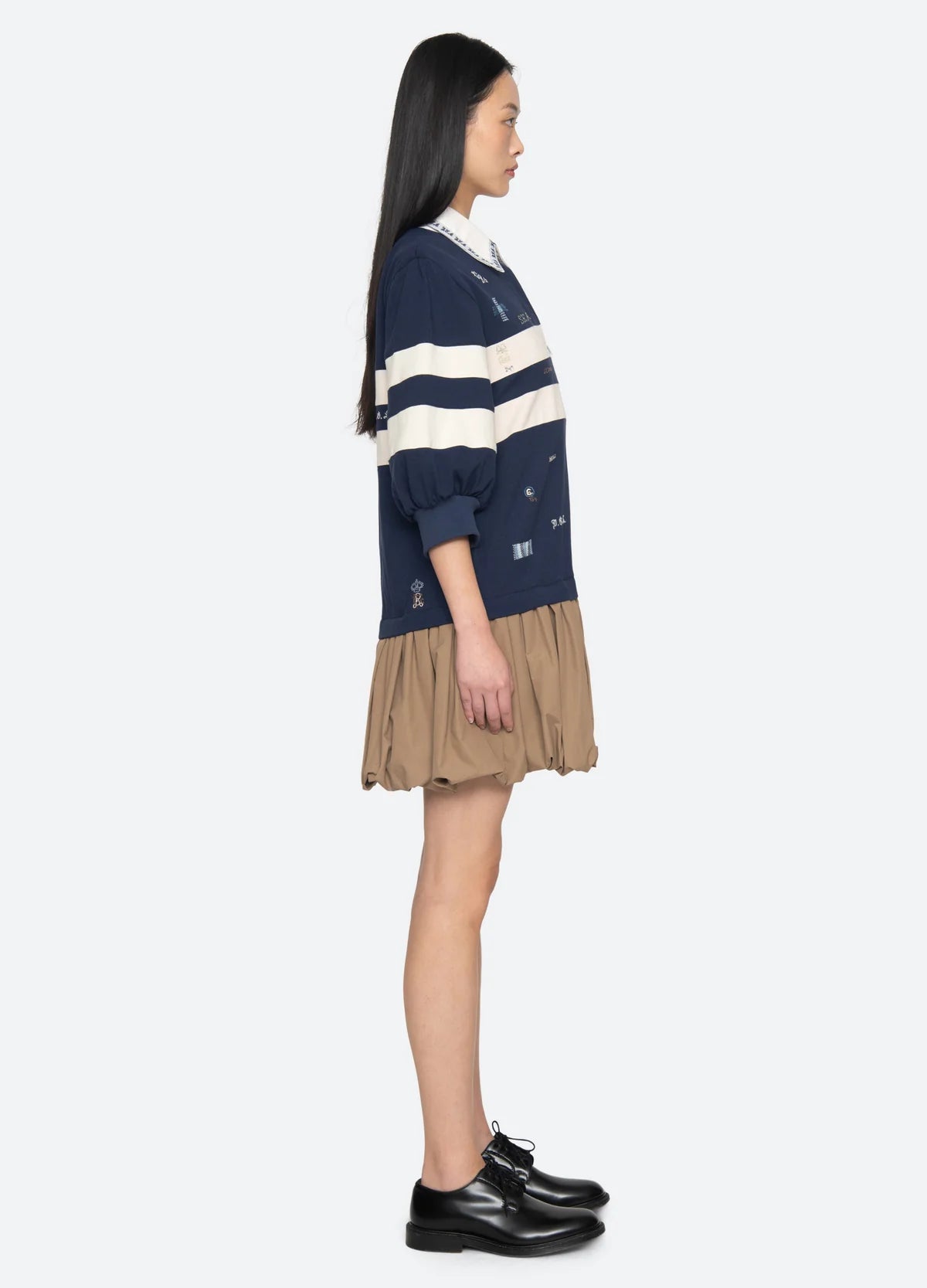 A woman stands in profile wearing the Sea New York Claira Rugby Dress—navy blue with white stripes and a beige bubble hem—paired with black shoes, set against a plain light background.