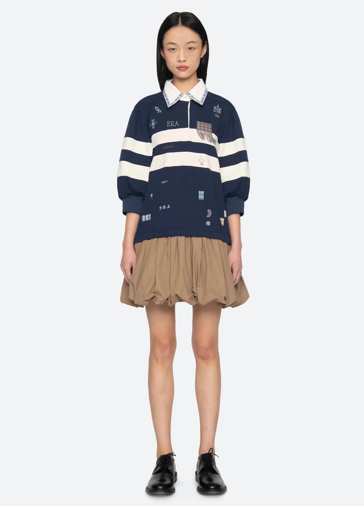 A woman stands facing forward, wearing the Sea New York Claira Rugby Dress—a navy blue piece with white stripes and embroidered patches—paired with black oxford shoes.