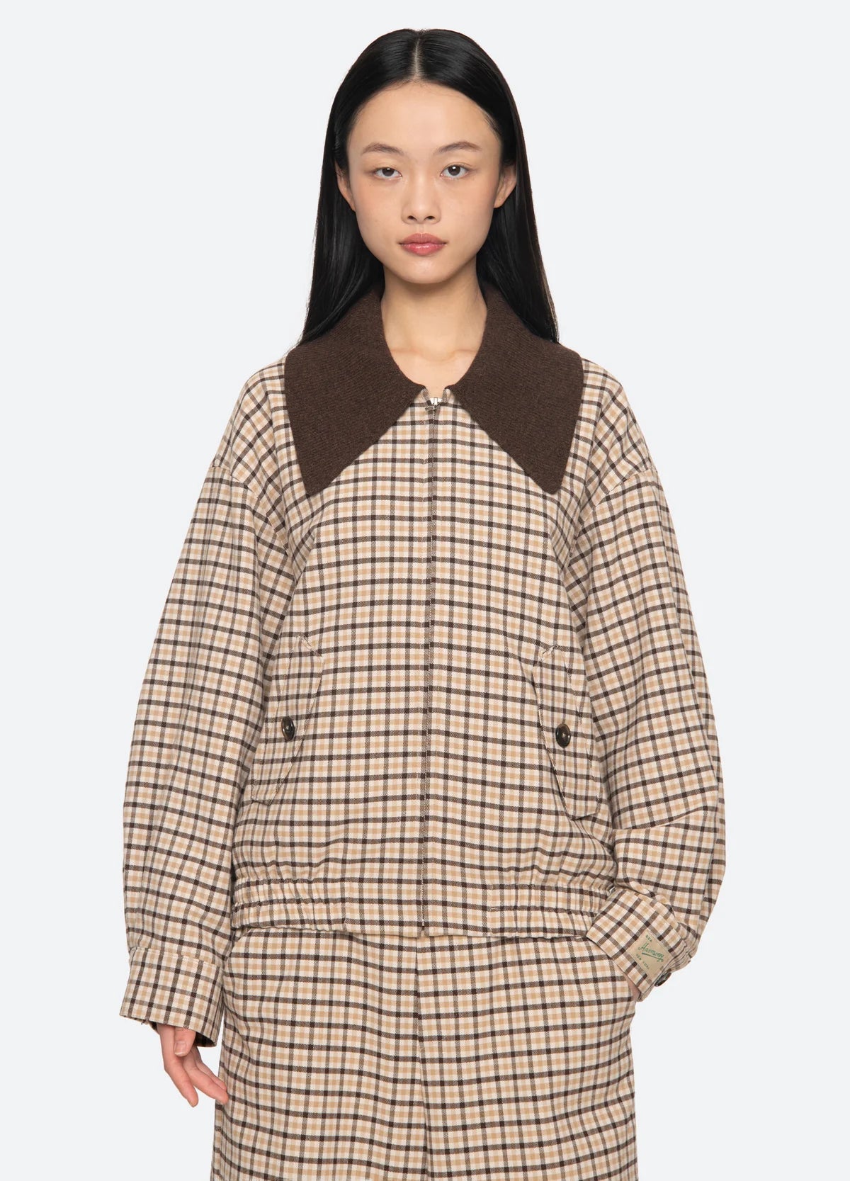 A person with long black hair models the Sea New York Savannah Plaid Jacket, which features an oversized ribbed-knit collar and a contemporary fit, standing against a plain light background.