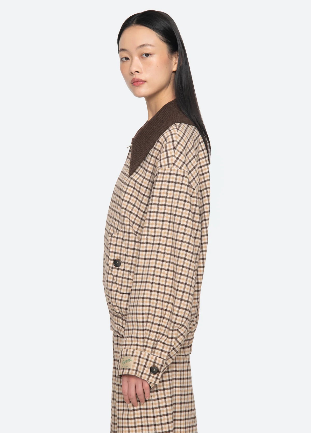 A woman with long, straight black hair poses in profile against a light gray background, wearing the Sea New York Savannah Plaid Jacket with a contemporary fit, matching pants, and an oversized ribbed-knit collar.