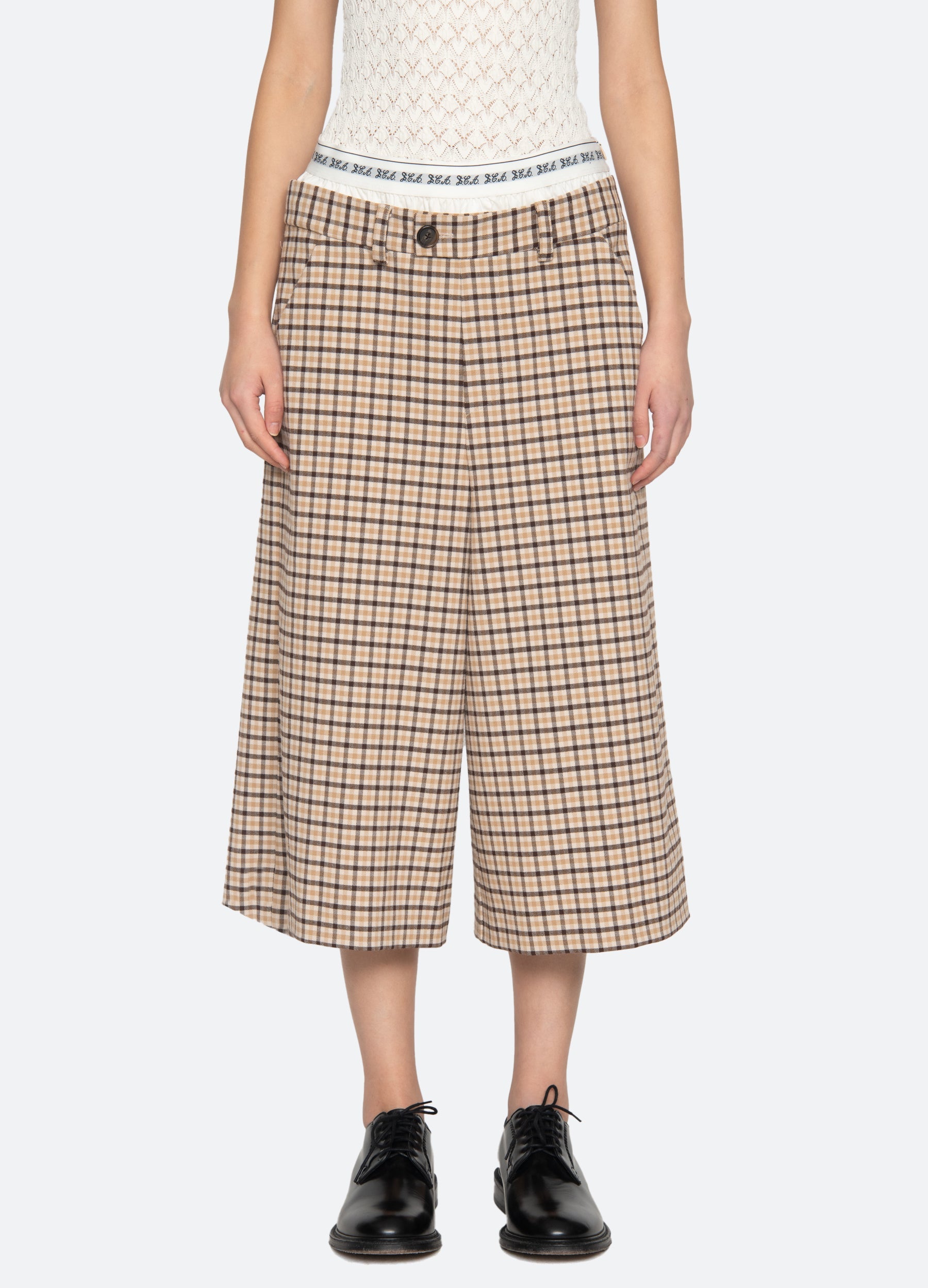 A person wears Sea New York's Savannah Plaid Culotte with a wide-leg fit, a white patterned sleeveless top, black lace-up shoes, and a visible boxer-style waistband featuring text, set against a plain light gray background.