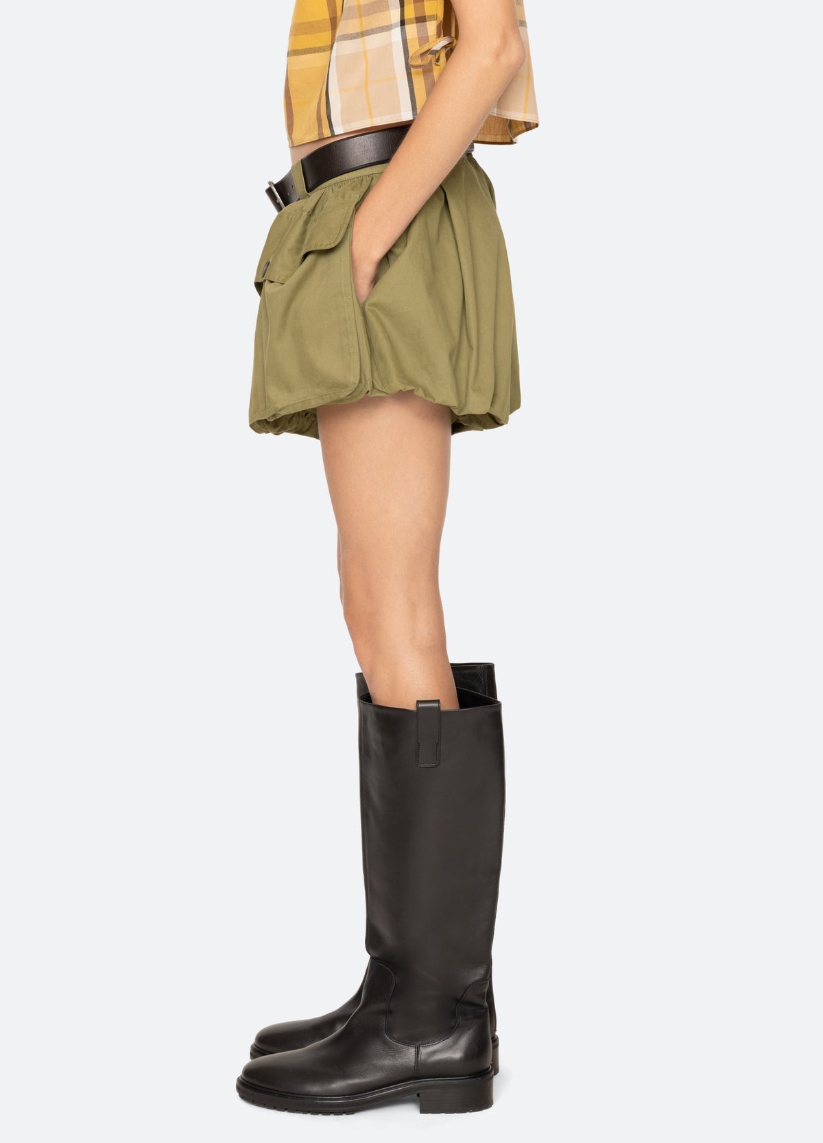A person in profile wears the Sea New York Andrea Bubble Short—olive green with oversized front pockets and removable leather belt—paired with a plaid shirt and knee-high black boots against a plain light background.