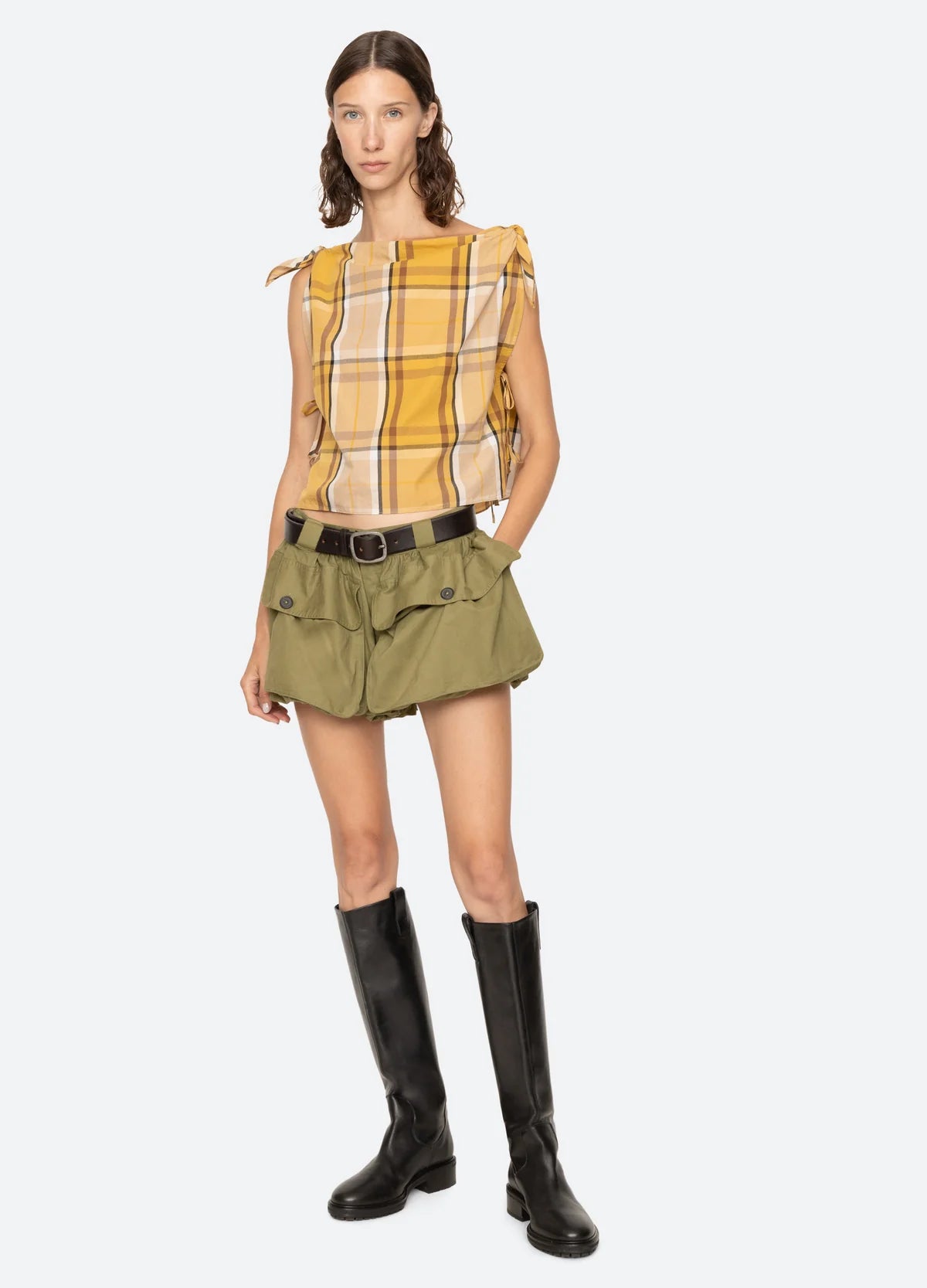 A woman wears a yellow plaid sleeveless top with shoulder ties, Sea New York's Andrea Bubble Short in olive green featuring oversized front pockets and a black belt, paired with knee-high black boots against a light backdrop.