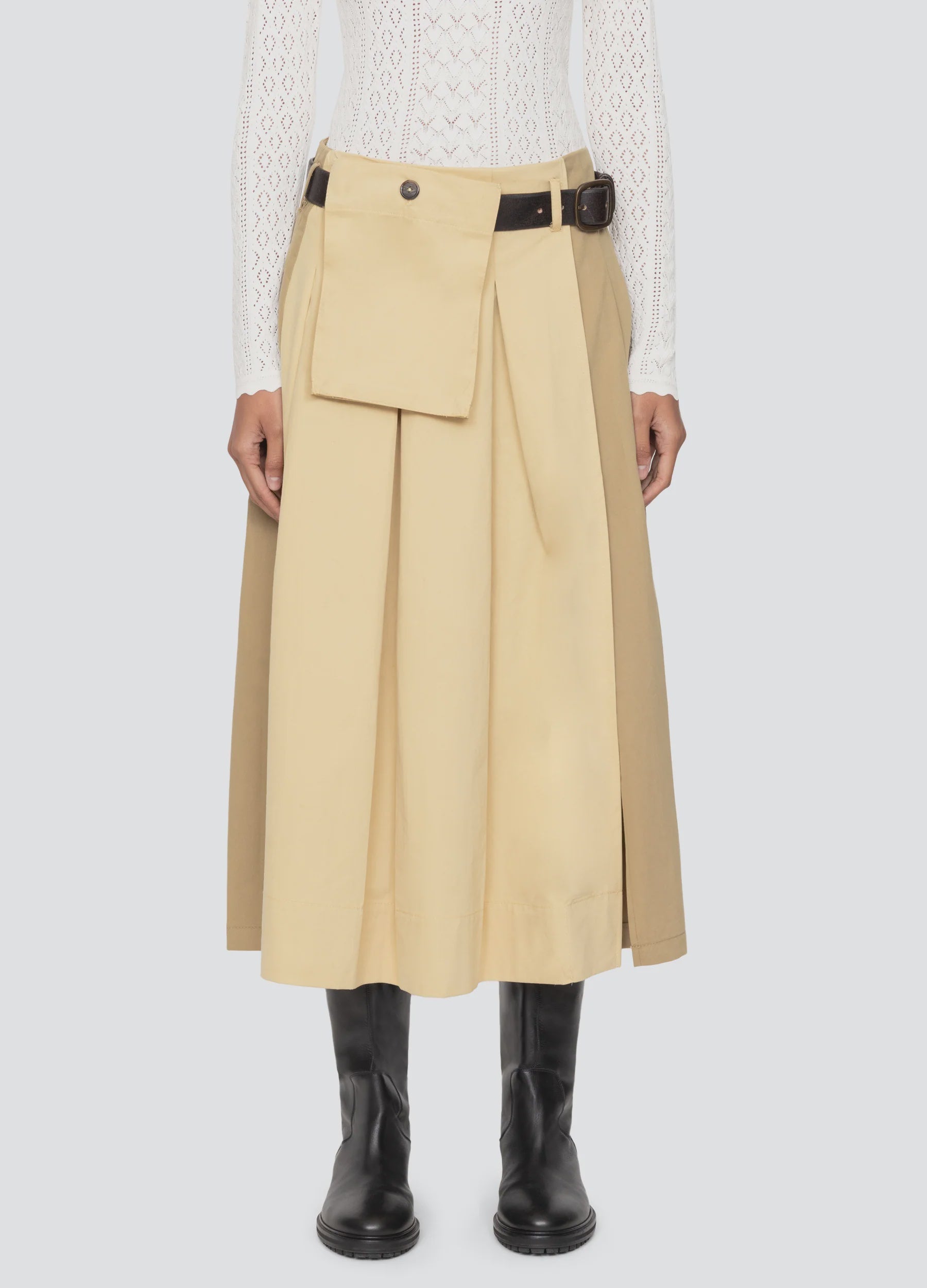 A person wearing the Sea New York Andrea Workwear Skirt, featuring a pleated midi design with a front pocket flap and leather belt, paired with a white textured long-sleeve top and black knee-high boots.