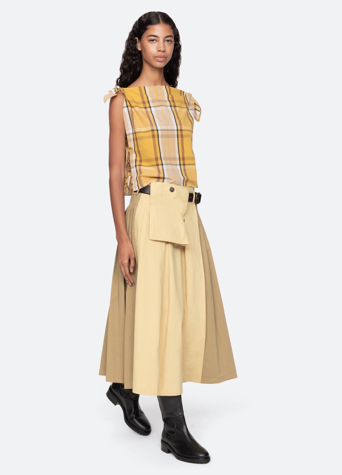 A woman in a yellow plaid sleeveless top, beige and tan Sea New York Andrea Workwear Skirt with leather belt and pouch, and black ankle boots stands against a plain light background.