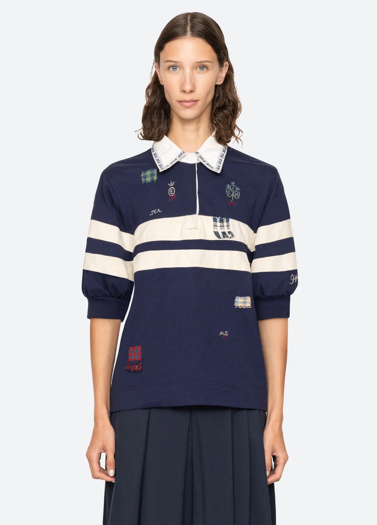 A woman with wavy brown hair wears the Claira Rugby Shirt by Sea New York—navy blue with white stripes, embroidered patches, and a crisp white collar—paired with a dark pleated skirt, standing against a plain light background.