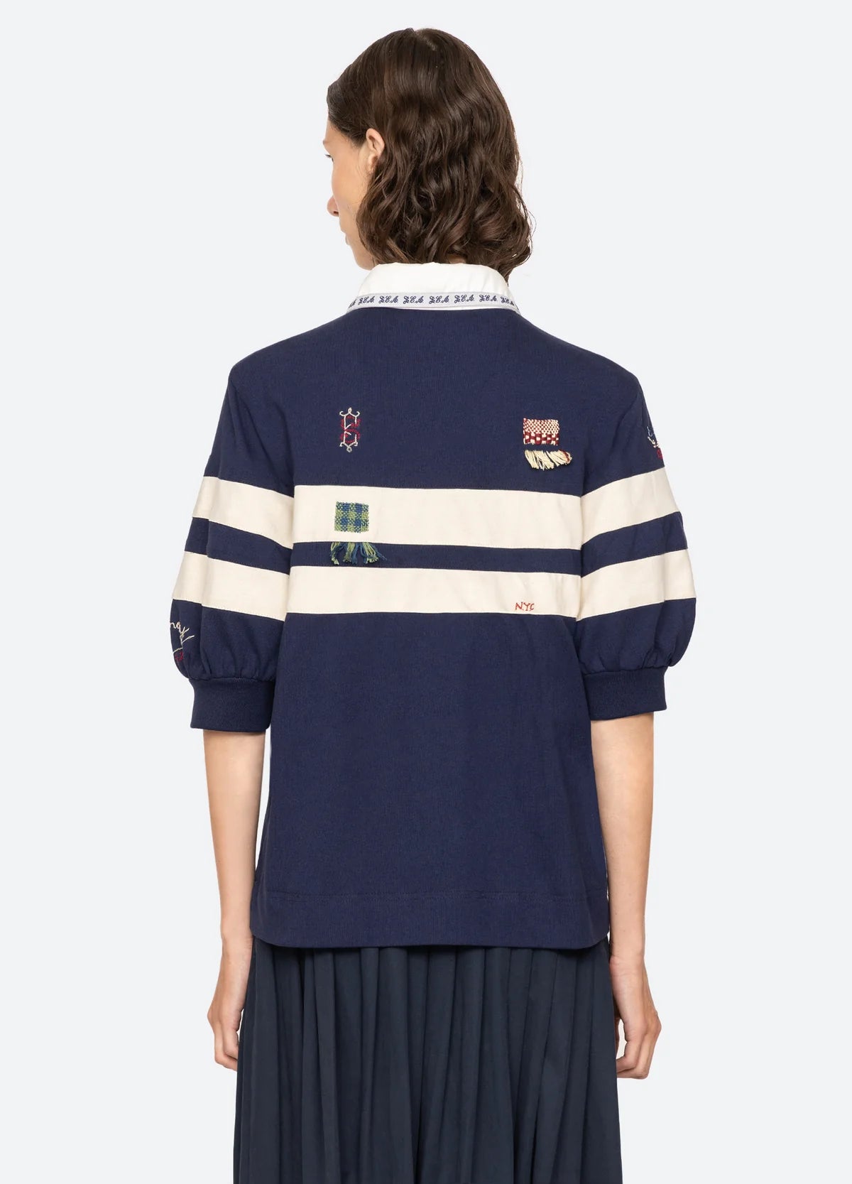 A person with short brown hair is seen from the back, wearing the Sea New York Claira Rugby Shirt—navy blue with white stripes, embroidered patches, and a white collar—paired with a dark pleated skirt.