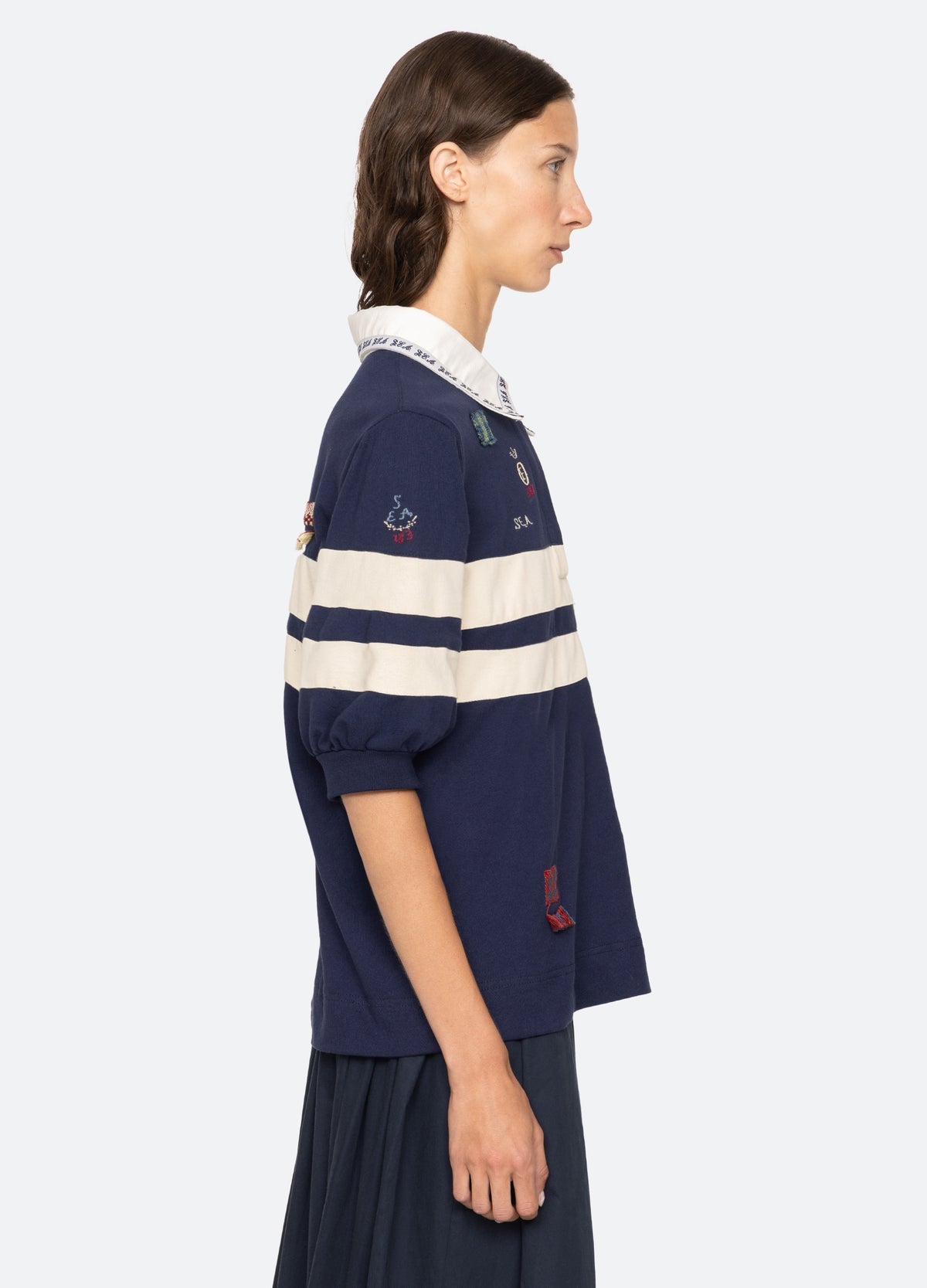 A woman with brown hair in a low ponytail models the Sea New York Claira Rugby Shirt—navy blue with white horizontal stripes and embroidered patches—standing in profile against a plain light background.
