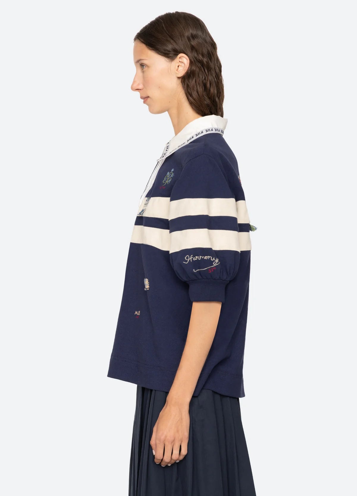 A woman with wavy brown hair stands in profile wearing the Sea New York Claira Rugby Shirt, featuring navy blue and white colors, embroidered details, and a pleated navy skirt against a plain light background.
