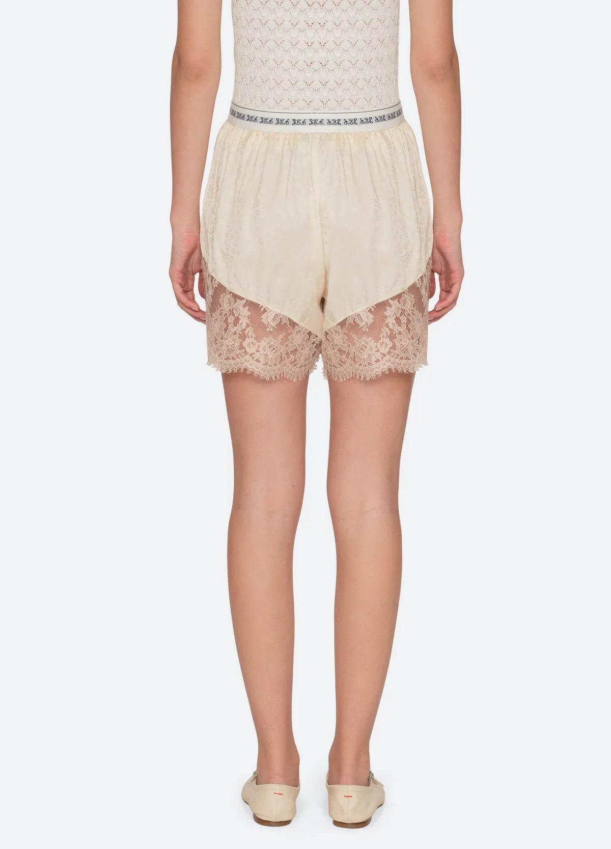 A person faces away wearing Ethel Solid Shorts by Sea New York in cream, featuring a lace hem and patterned elastic waistband, paired with cream slip-on shoes and a textured sleeveless top, set against a plain white background.