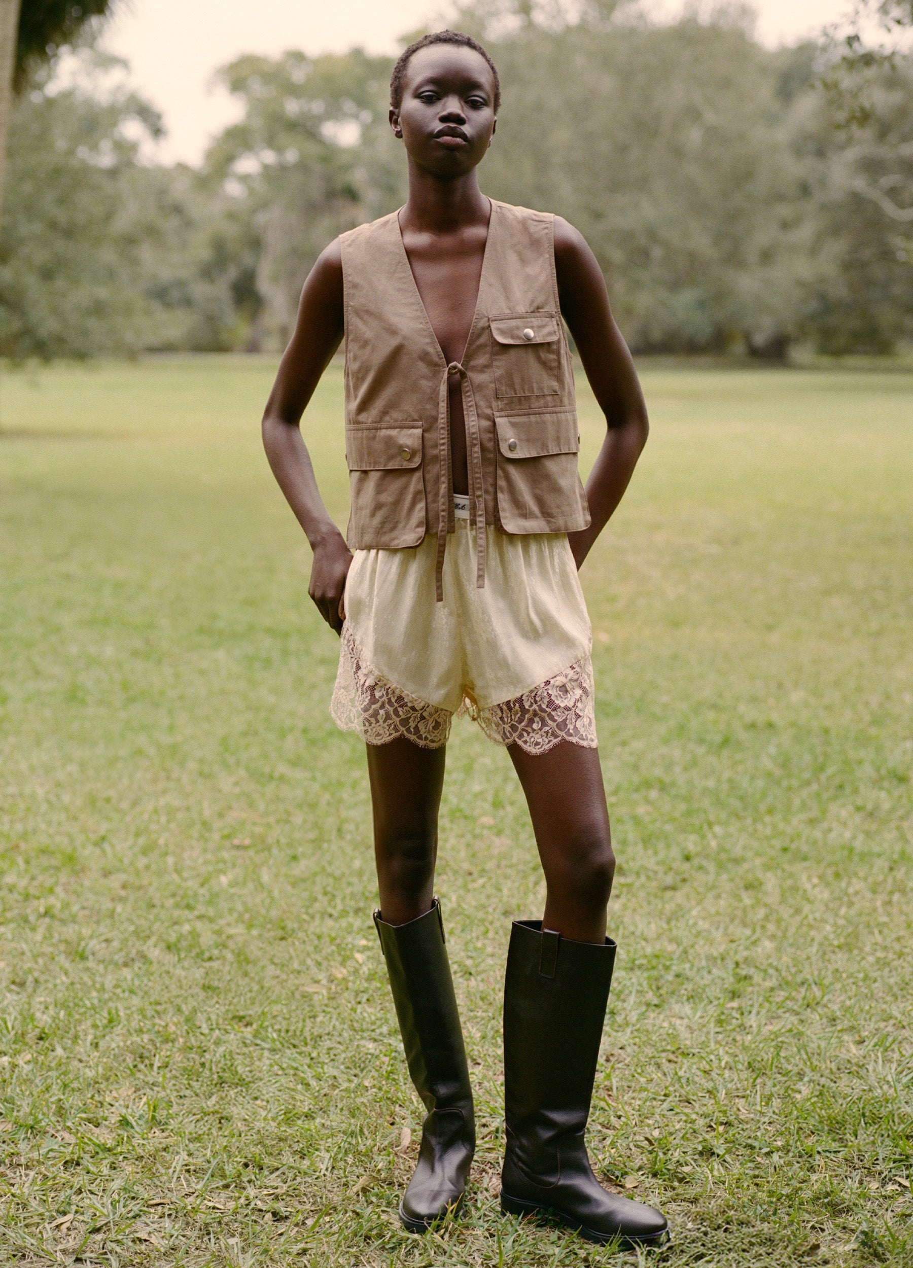 A person stands on grass outdoors, wearing the Ethel Solid Shorts by Sea New York, a tan sleeveless vest, and black knee-high boots. Trees are visible in the blurred background.