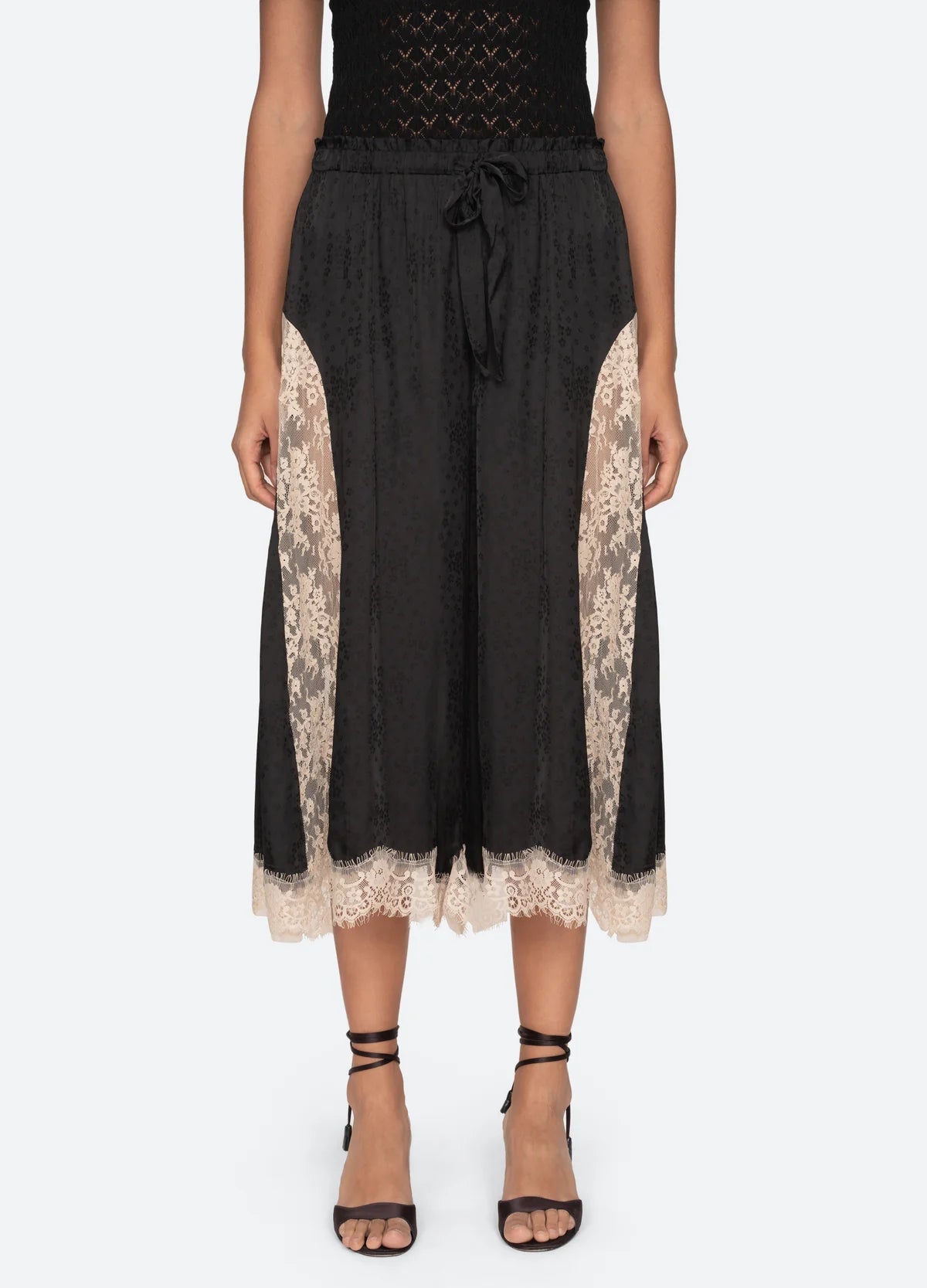 A person wears the Ethel Solid Pants by Sea New York—black culottes with beige lace trim, adjustable waistband—paired with a black top and strappy heels, standing against a light background.