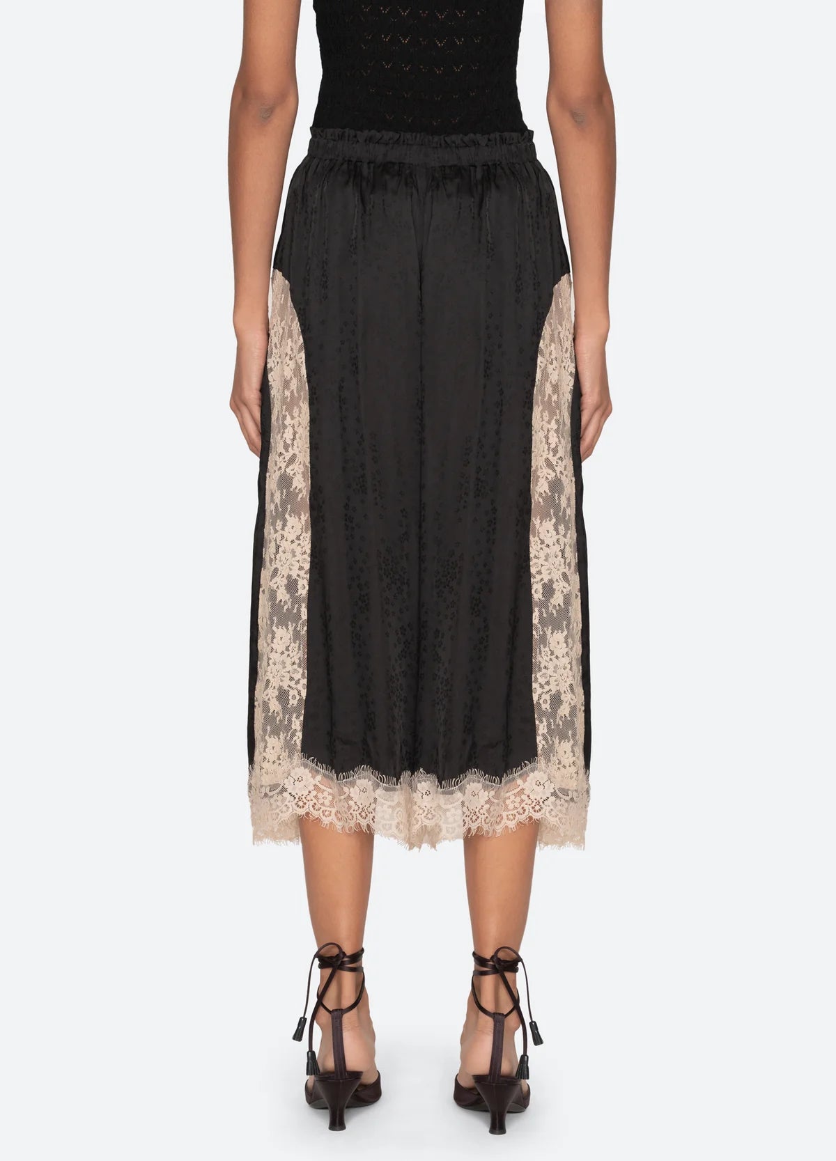 A woman is seen from the waist down wearing Sea New York’s Ethel Solid Pants—black midi-length pants featuring beige lace panels on the sides and hem, styled with black strappy heels.