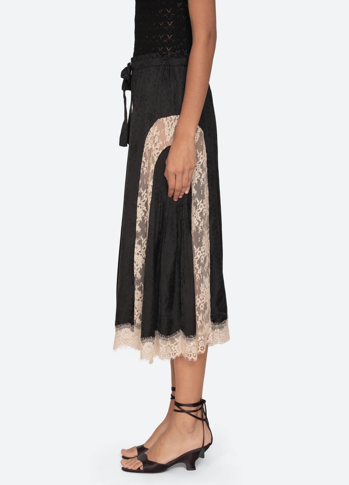 A woman stands in profile against a light background, wearing Sea New York's Ethel Solid Pants with a sleeveless black top and relaxed-fit black low-heeled sandals tied at the ankle.