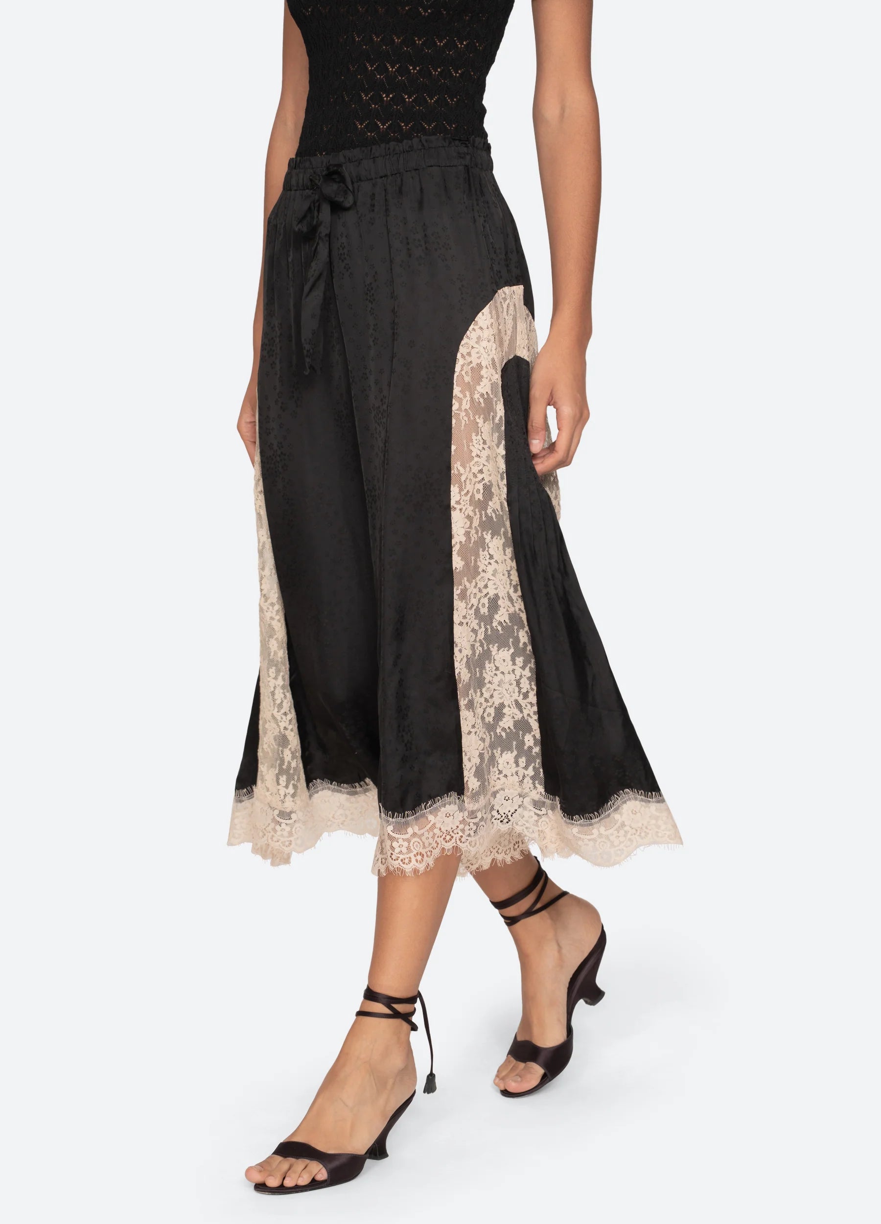 A woman wears the Ethel Solid Pants by Sea New York with a black sleeveless top and black strappy heeled sandals.