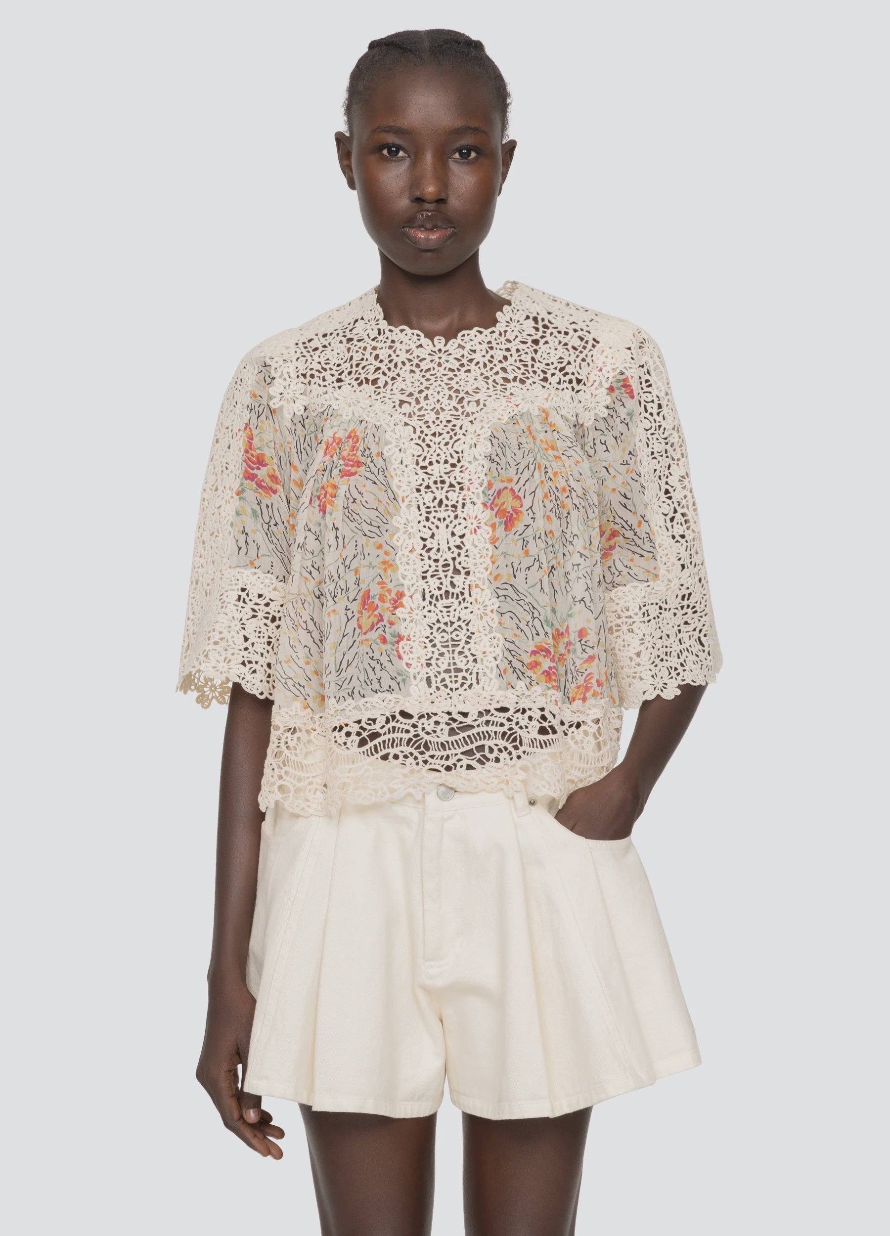 A woman wears the Sea New York Luisa Print SS Top—a sheer, cream lace blouse with floral embroidery—and loose, high-waisted ivory shorts. She stands with one hand in her pocket against a plain light background.