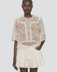 A woman wears the Sea New York Luisa Print SS Top—a sheer, cream lace blouse with floral embroidery—and loose, high-waisted ivory shorts. She stands with one hand in her pocket against a plain light background.